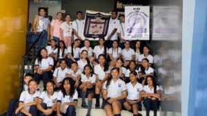 Grupo de estudiantes y docentes de la Institución Educativa Juan XXIII posando en unas escaleras dentro de un edificio. Los estudiantes, vestidos con uniforme blanco y azul, sostienen dos carteles con el nombre de la institución y mensajes educativos. El ambiente refleja unión y trabajo en equipo.