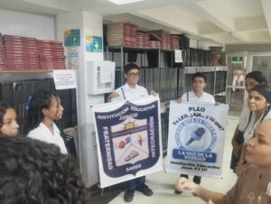 Grupo de estudiantes en uniforme escolar dentro de una sala con estanterías llenas de libros encuadernados. Dos estudiantes sostienen carteles de la Institución Educativa Juan XXIII con mensajes de fraternidad, saber e integración, y del noticiero estudiantil ‘La voz de la verdad’. El grupo conversa en círculo, mostrando interés en la actividad.