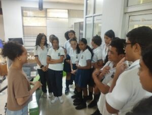 Un grupo de estudiantes en uniforme escolar escucha con atención a una mujer adulta que les habla en una sala del periódico El Meridiano. Los jóvenes están de pie en semicírculo, mostrando interés en la conversación.